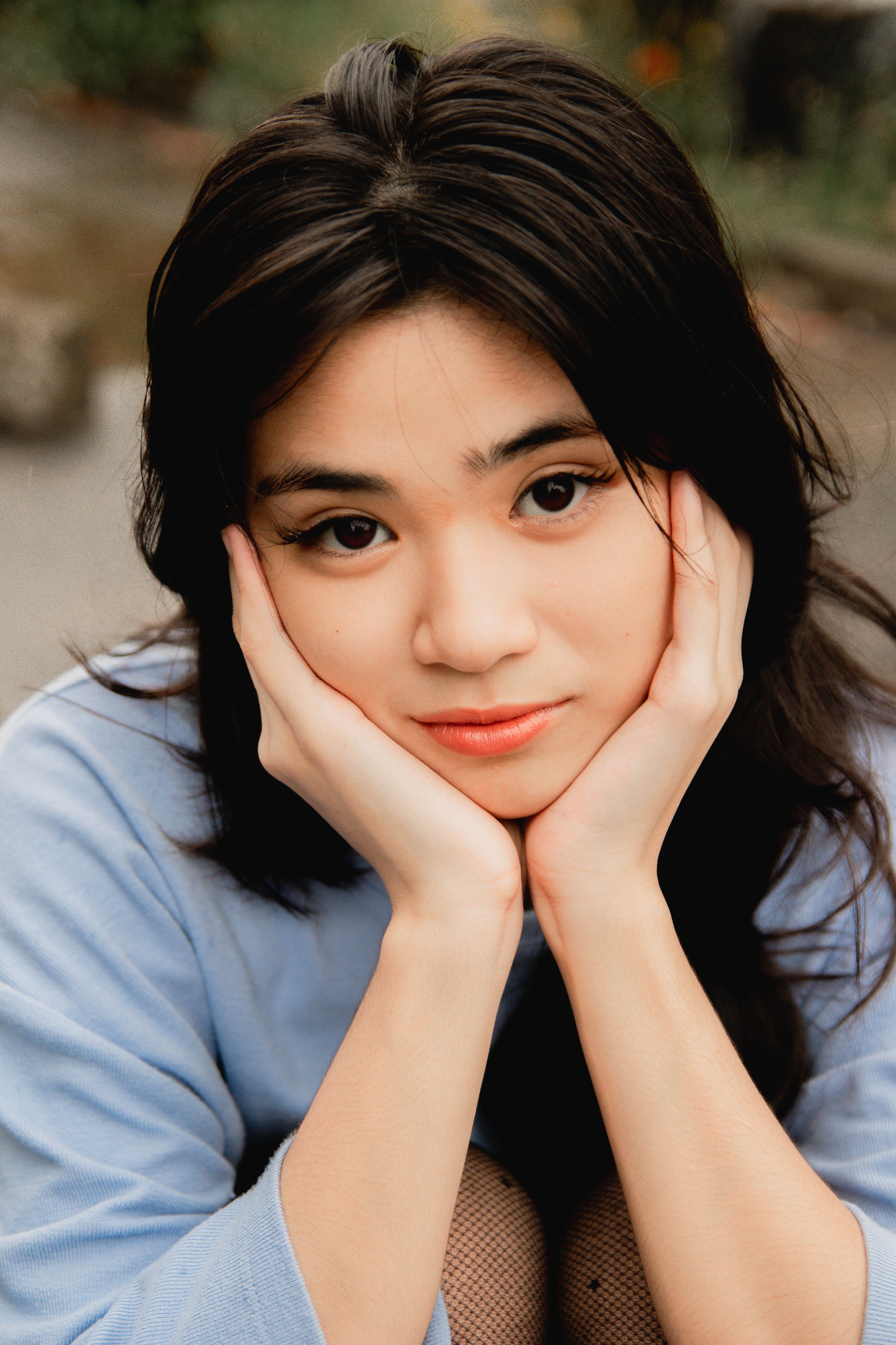 Close-up portrait of a young woman in a blue sweater, posing outdoors with hands on chin.