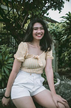 Cheerful woman in a yellow gingham top posing outdoors surrounded by greenery.