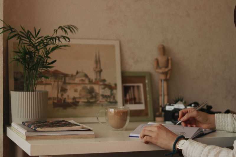 A person writing in a notebook at a calm home desk with coffee and a plant