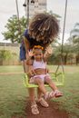 Anonymous mom with Afro hairstyle leaning forward above cheerful ethnic girl while having fun on swing in urban park