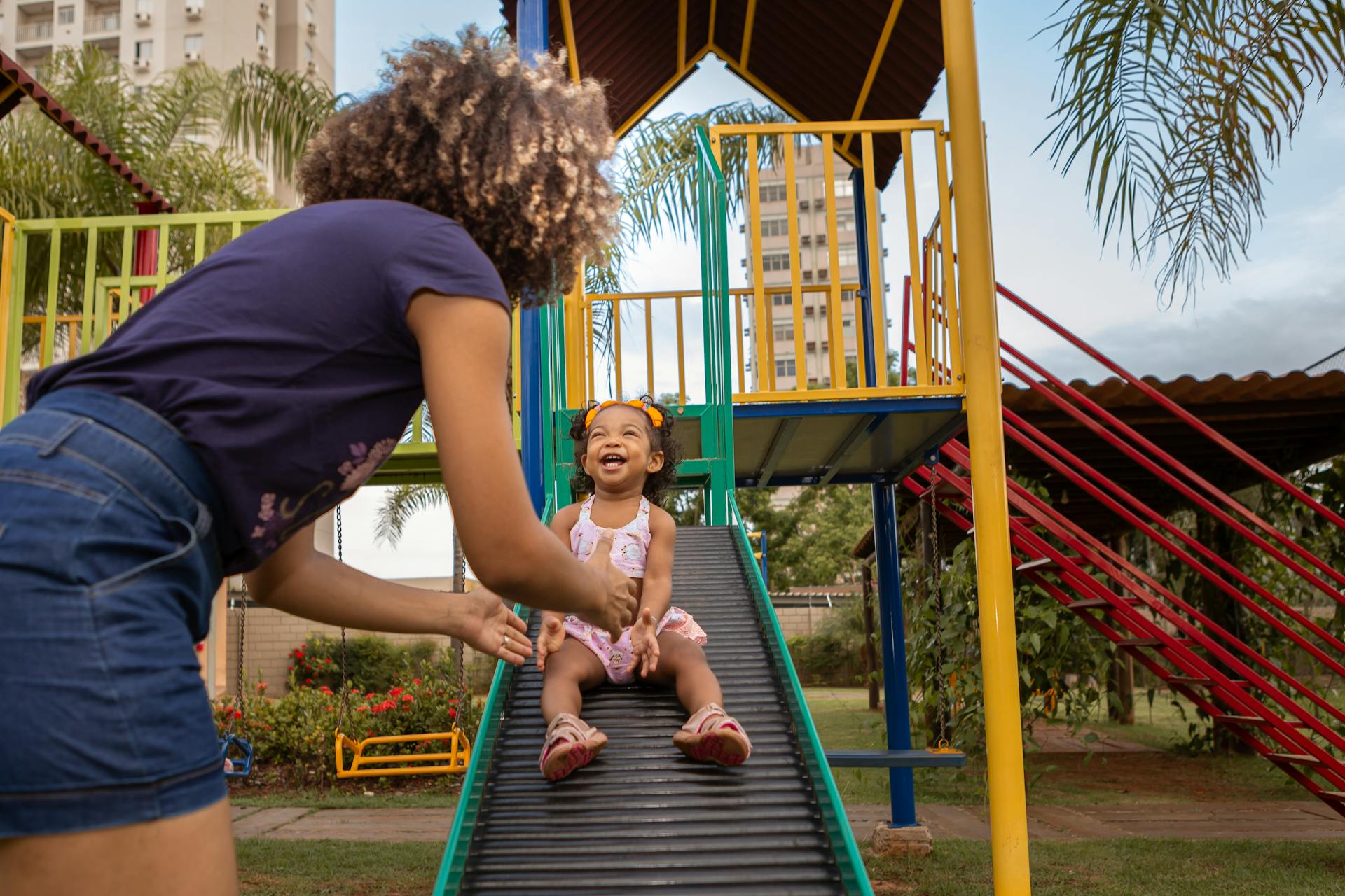 A young girl with afro hair joyfully slides down, playing in a vibrant outdoor playground.