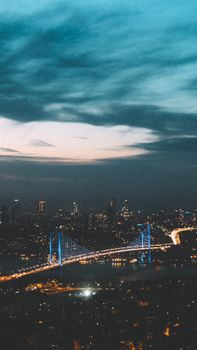 Long bridge with glowing lights crossing river in modern city with illuminated buildings against cloudy sky at dark night time