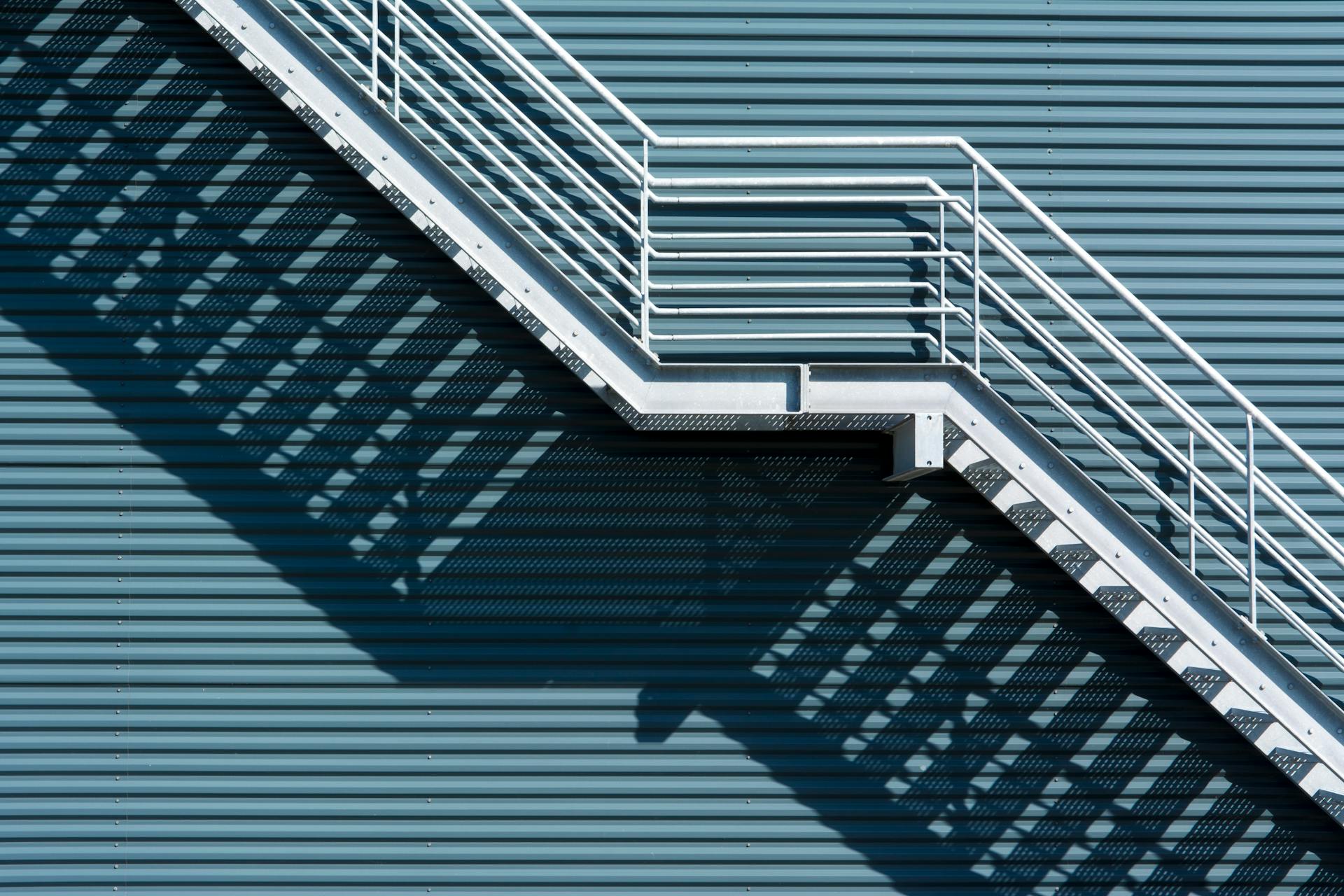 An outdoor metal staircase casting geometric shadows on a corrugated blue wall.