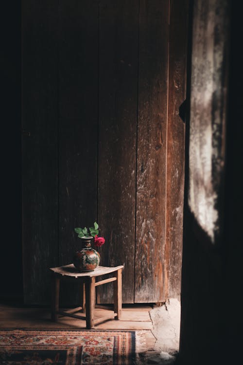 Old chair with flower in vase placed near carpet in shabby room with wooden walls in sunlight
