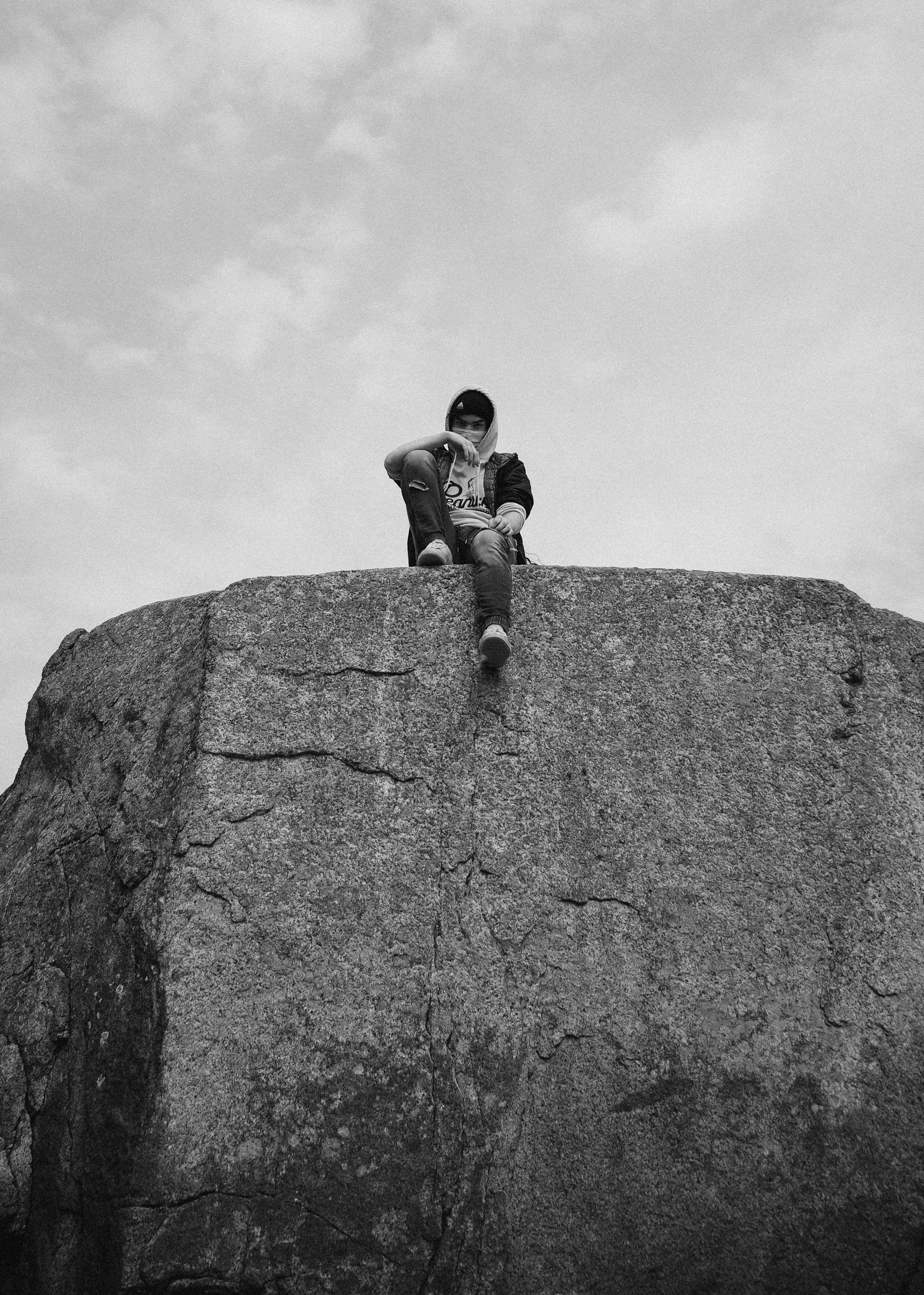 A person sitting atop a large rock wearing a hoodie in a grayscale outdoor setting.