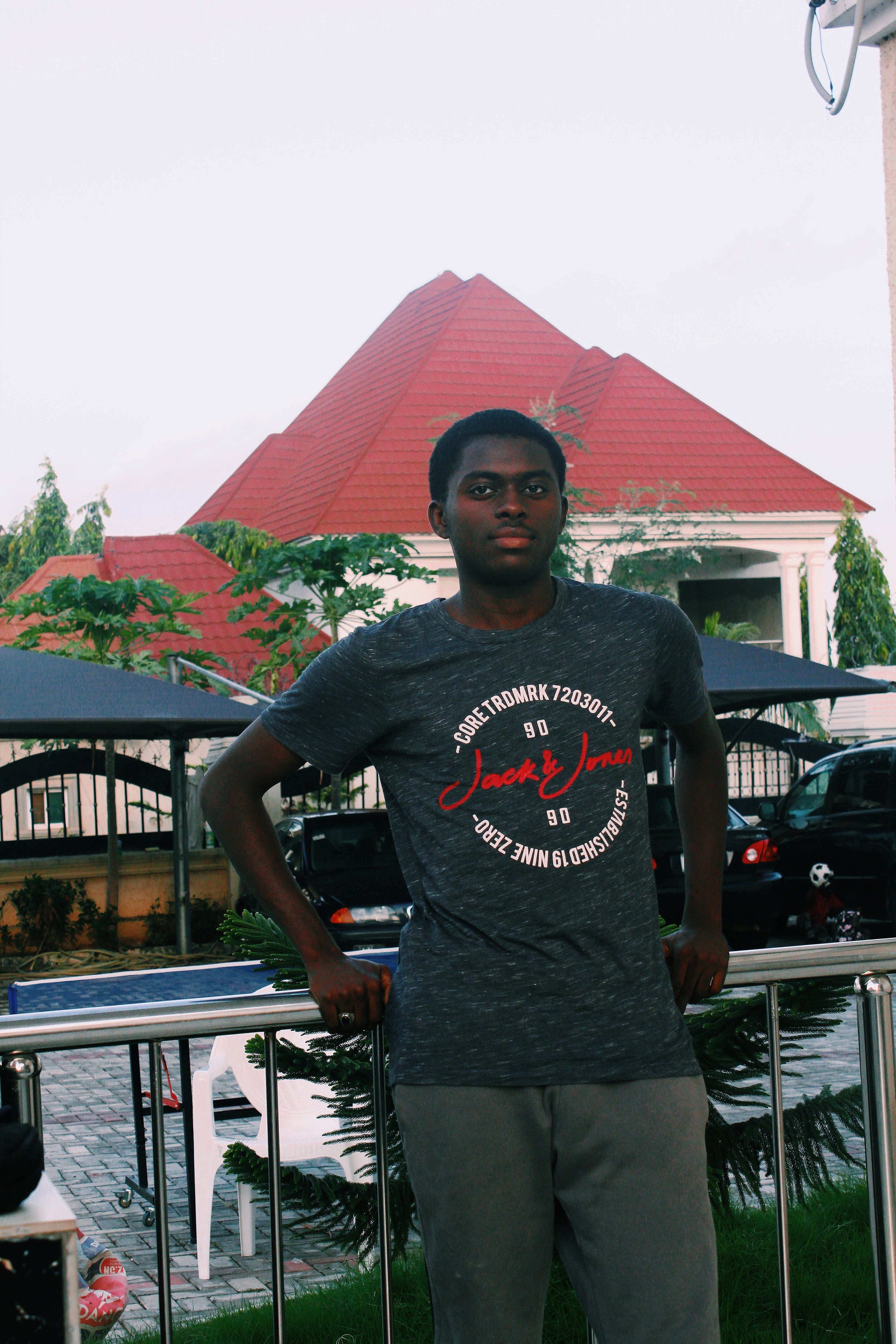 Serious African American male in t shirt and pants standing near metal railing at backyard against parked cars and looking at camera
