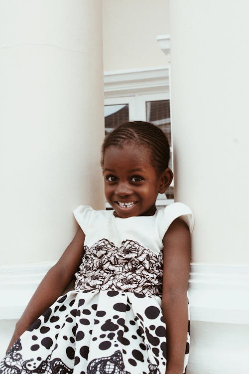Adorable little African American girl wearing dress smiling brightly while sitting between white columns and looking at camera