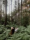Back view of unrecognizable female with long hair standing among green plants in forest with tall tress in summer