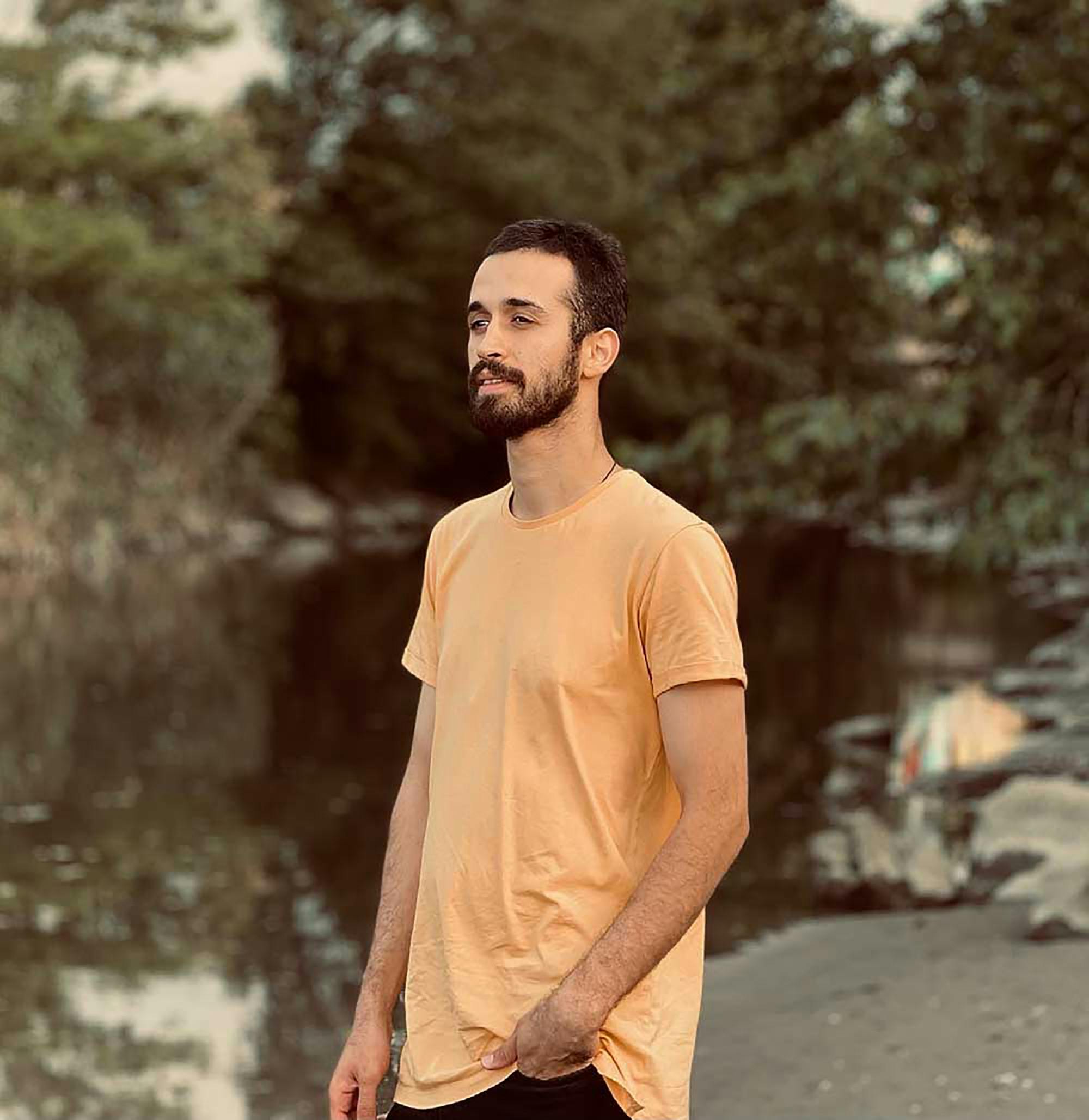 Young man in a yellow shirt standing near a river in a serene outdoor setting.