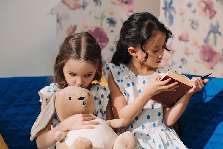 Young Girls Sitting Together At The Bed While Reading A Book And Holding A Plush Toy