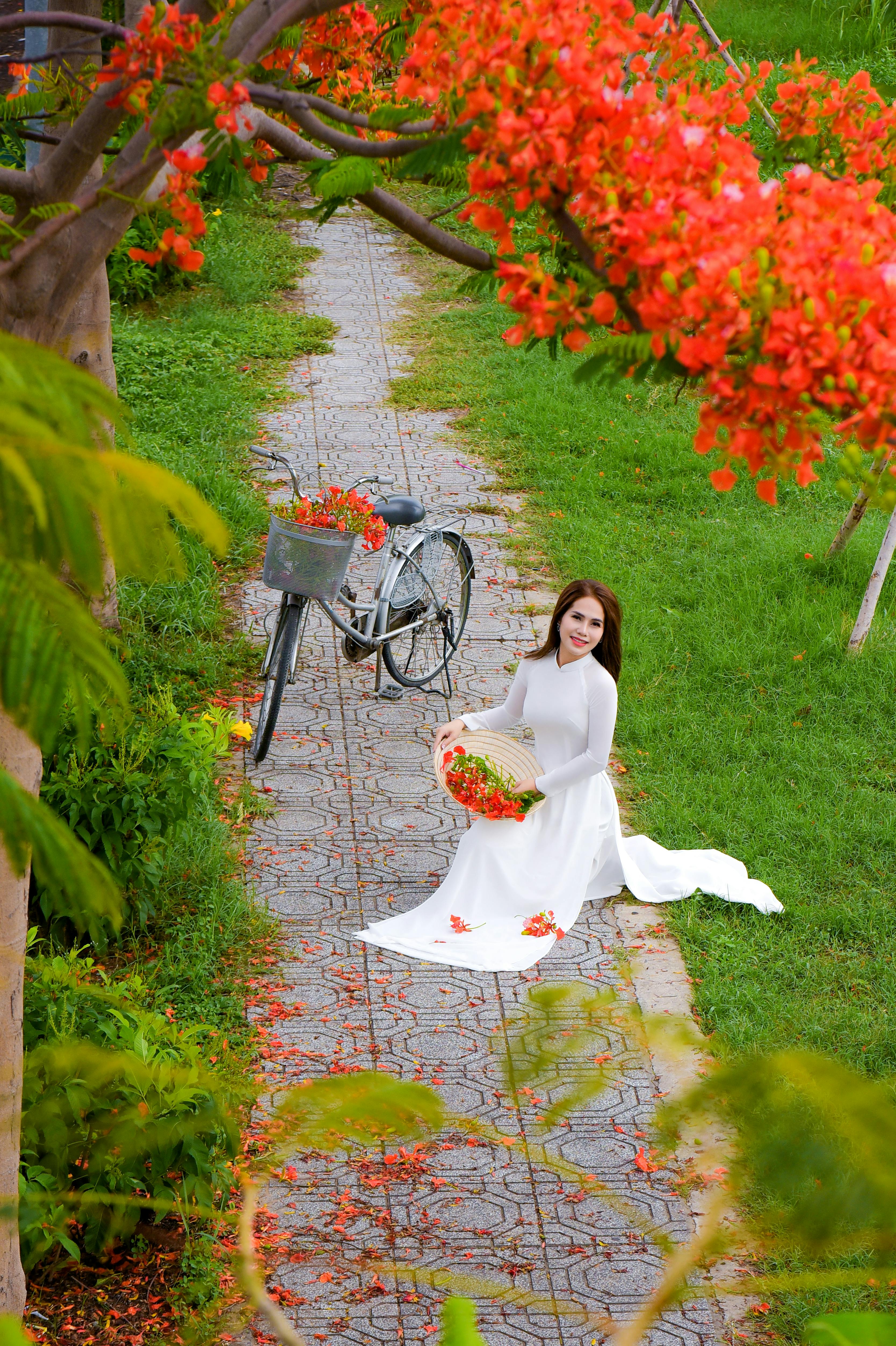 Asian woman in traditional Ao Dai kneeling by bicycle with vibrant flowers on a garden pathway.