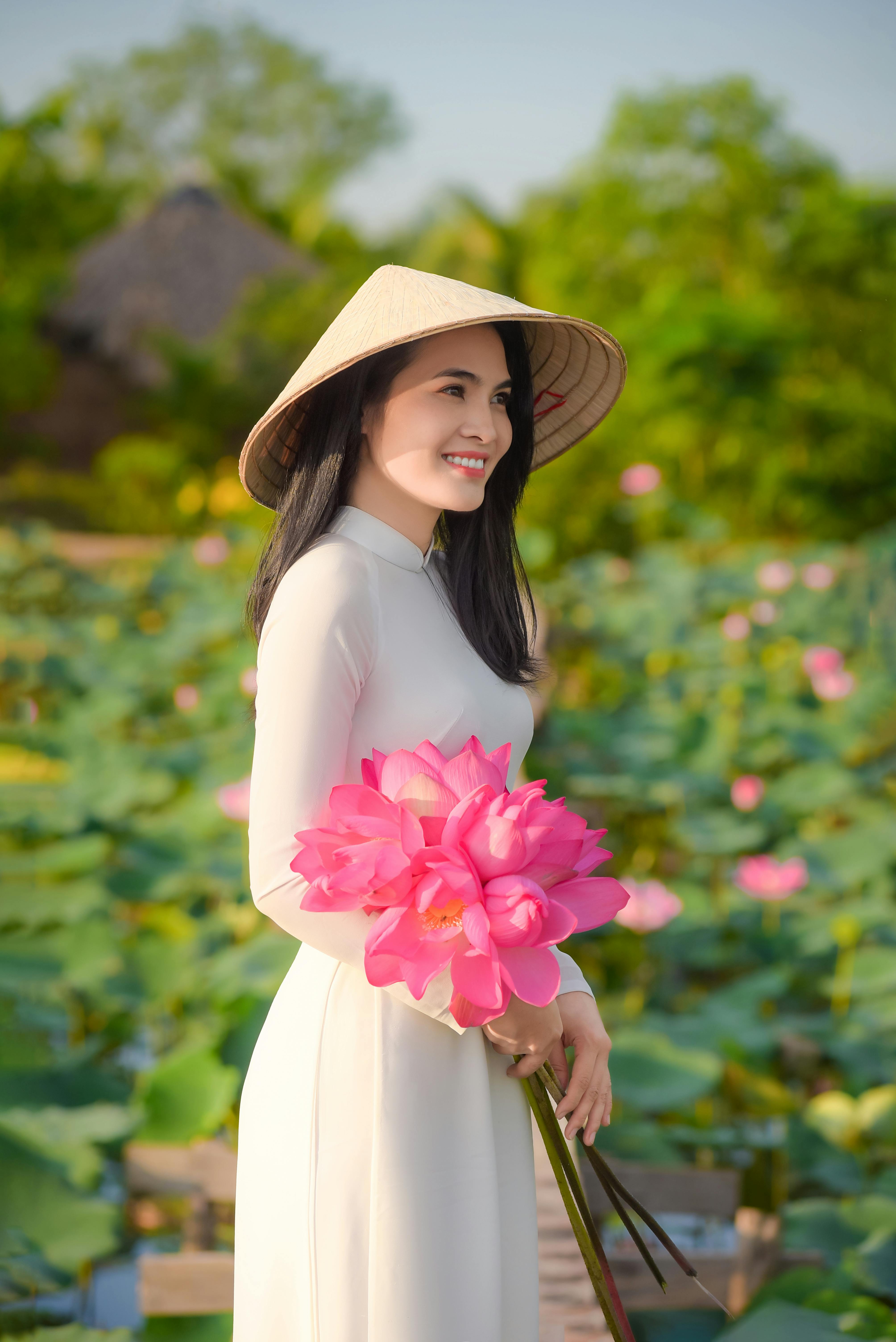 Asian woman in white traditional dress and conical hat holding pink lotus flowers, smiling outdoors.