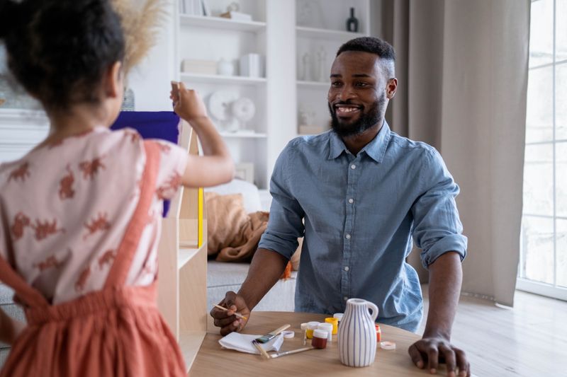 A smiling Black father sits at a table with his young daughter as they paint together at home.