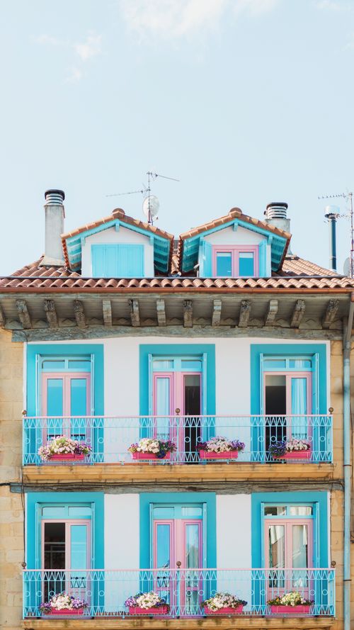 Free Colorful residential building with blue window frames and flower-filled balconies against a clear sky. Stock Photo