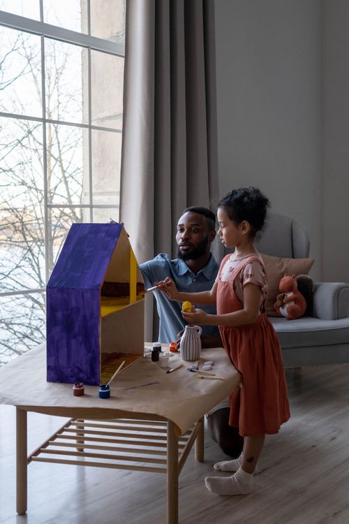 Free A father and daughter enjoy painting a dollhouse indoors, spending quality family time together. Stock Photo