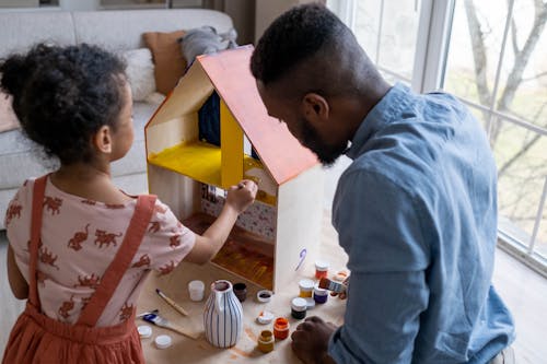 Back view of African American father and little daughter painting with gouache colors wooden toy house for dolls