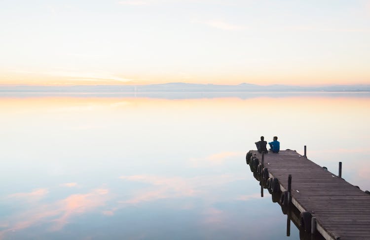 Two Men Sitting At The Edge Of A Lakeshore Jetty At Dusk