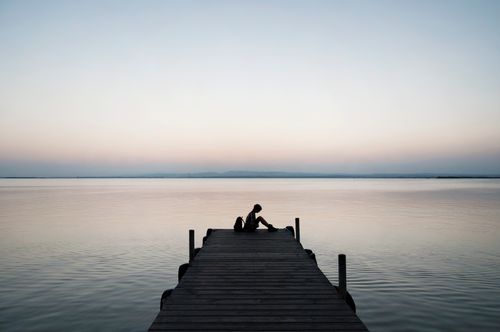 Free Silhouette of a person sitting on a dock at sunset by a calm sea. Stock Photo