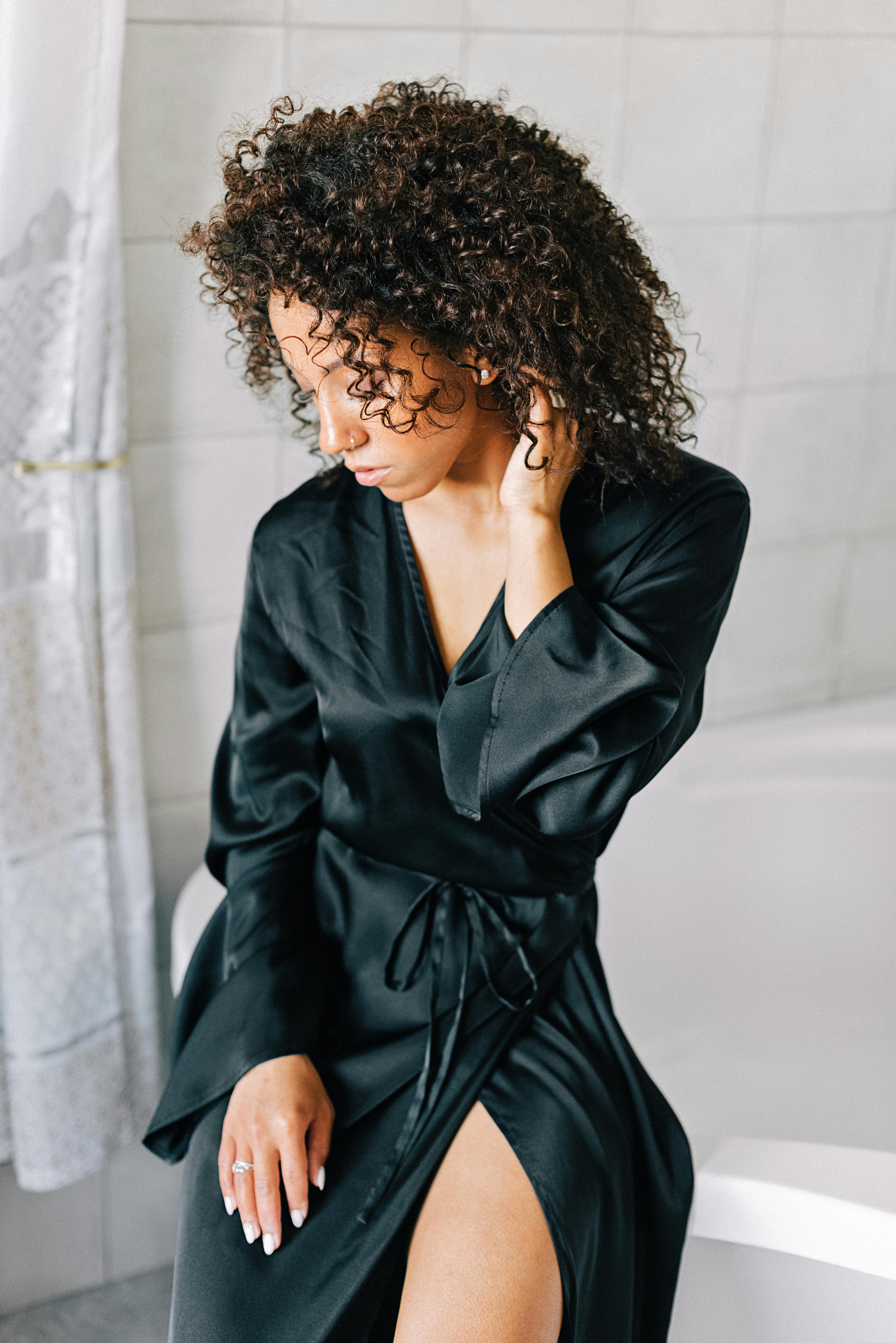 Stylish woman in black silk robe with curly hair, posing indoors next to a bathtub.