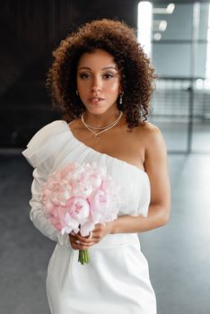 A stunning bride in a stylish white dress holding a pink bouquet, captured indoors with a modern flair.