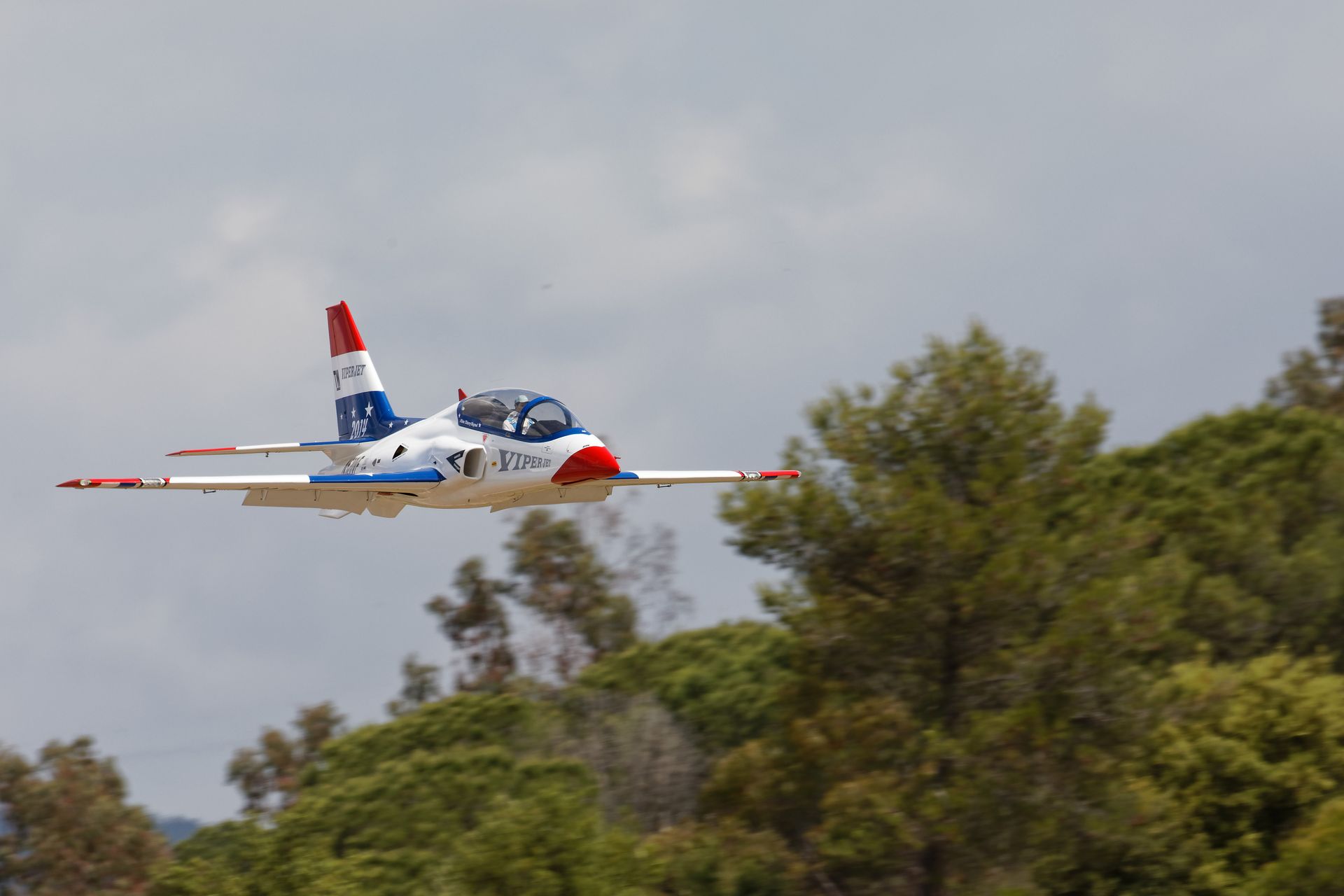 Colorful jet aircraft flying low over lush green trees on a clear day.