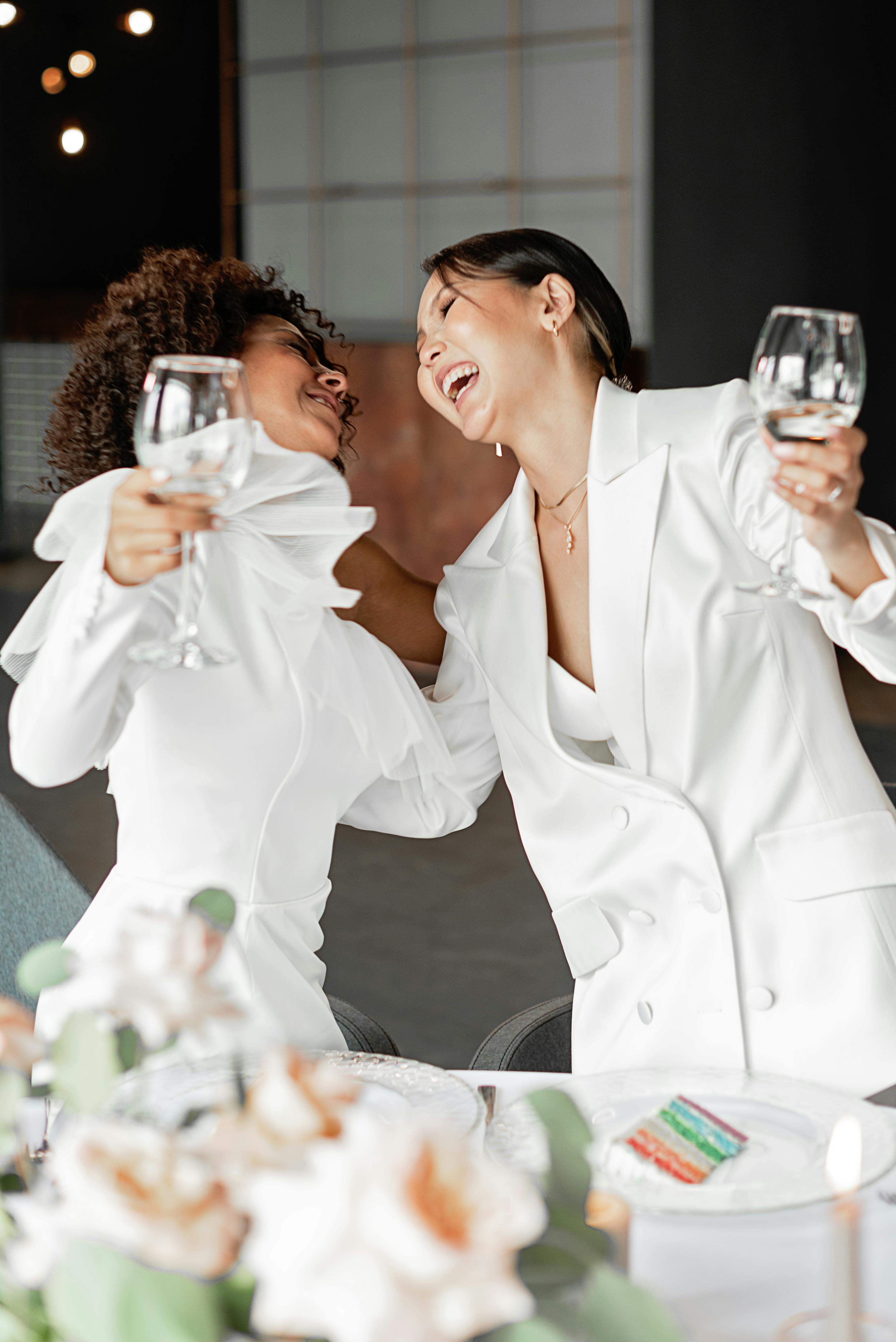 Two happy women in white attire celebrating with wine glasses, capturing joy and togetherness.