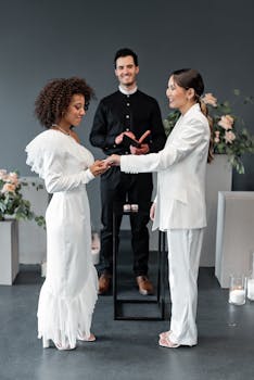 Two women exchange rings in a modern indoor wedding ceremony, officiated by a priest.