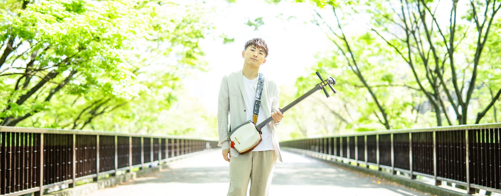 An Asian man standing on a sunlit bridge, holding a shamisen, surrounded by vibrant green trees.