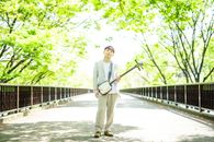An Asian man standing on a sunlit bridge, holding a shamisen, surrounded by vibrant green trees.