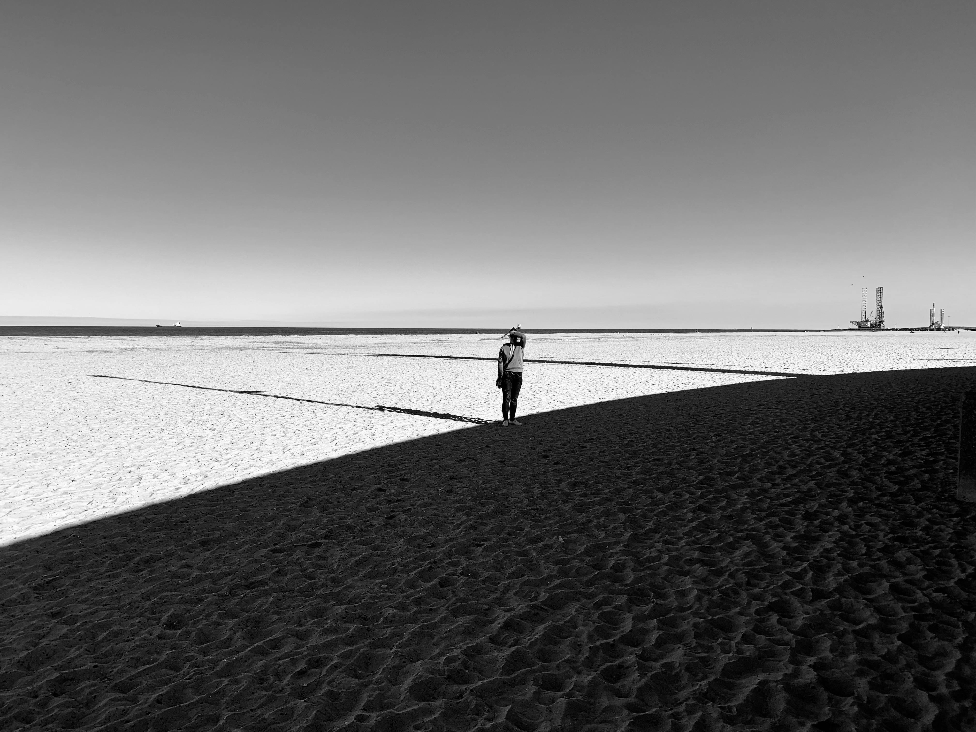 A lone person walks on a beach under dramatic light and shadow in black and white.