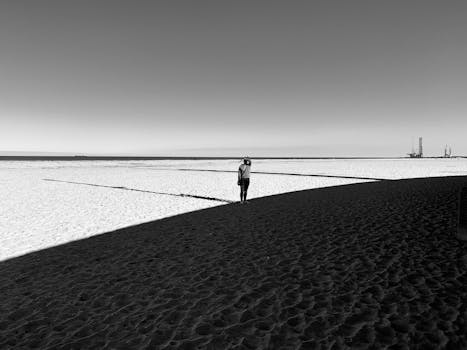 A lone person walks on a beach under dramatic light and shadow in black and white.
