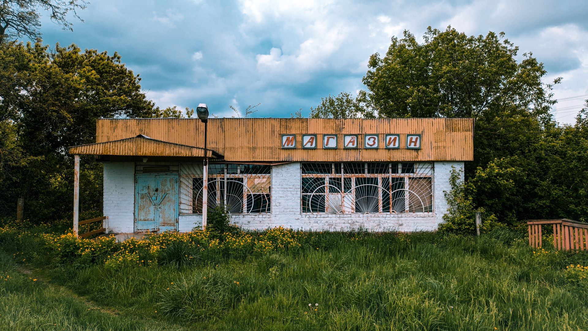 An abandoned rural store with overgrown grass and trees, indicative of nature reclaiming an old structure.