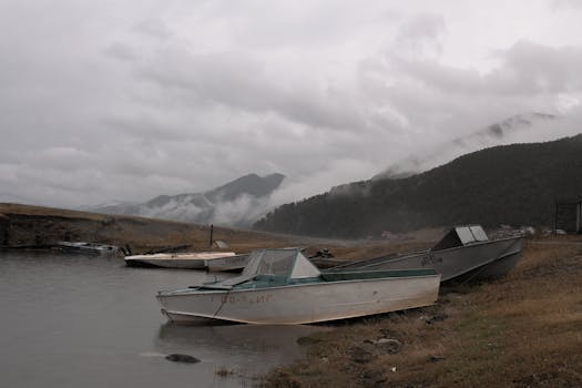 Several moored boats resting on a misty lakeshore against a backdrop of hills and overcast sky.
