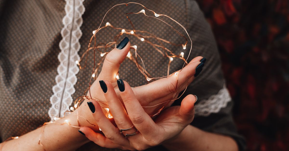 Close-up of hands with black nails holding glowing Christmas string lights in festive ambiance.