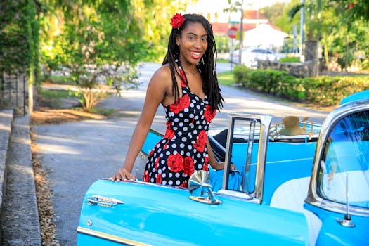 A cheerful woman in a floral dress stands beside a classic blue car on a sunny day.