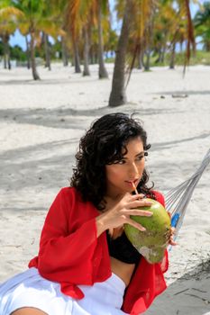 Woman in a red top relaxing on a tropical beach, sipping coconut water from a hammock.