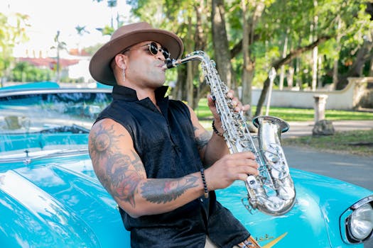 Musician with tattoos and hat plays saxophone near vintage blue car outdoors.