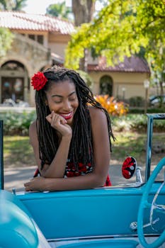 Cheerful woman in a vintage car surrounded by lush greenery on a sunny day.