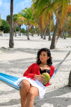 A woman enjoys a tropical drink in a hammock on a sunny beach, surrounded by palm trees.