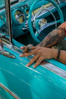 Close-up of a tattooed hand holding a cigar over a vintage teal convertible steering wheel.