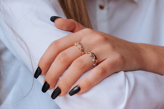 Close-up of hands with black nail polish and elegant rings against a white shirt backdrop.