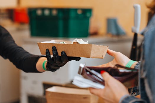 Person hands over food order in cardboard tray wearing black gloves in restaurant.