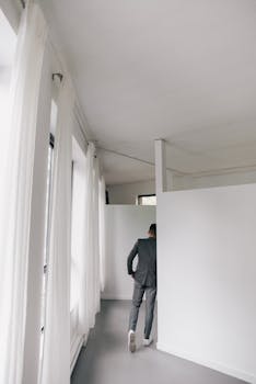 A man in a suit walking through a minimalist white hallway with natural light.