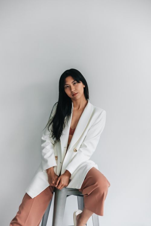 Free Portrait of an Asian woman in a white blazer sitting on a stool with a minimalistic backdrop. Stock Photo