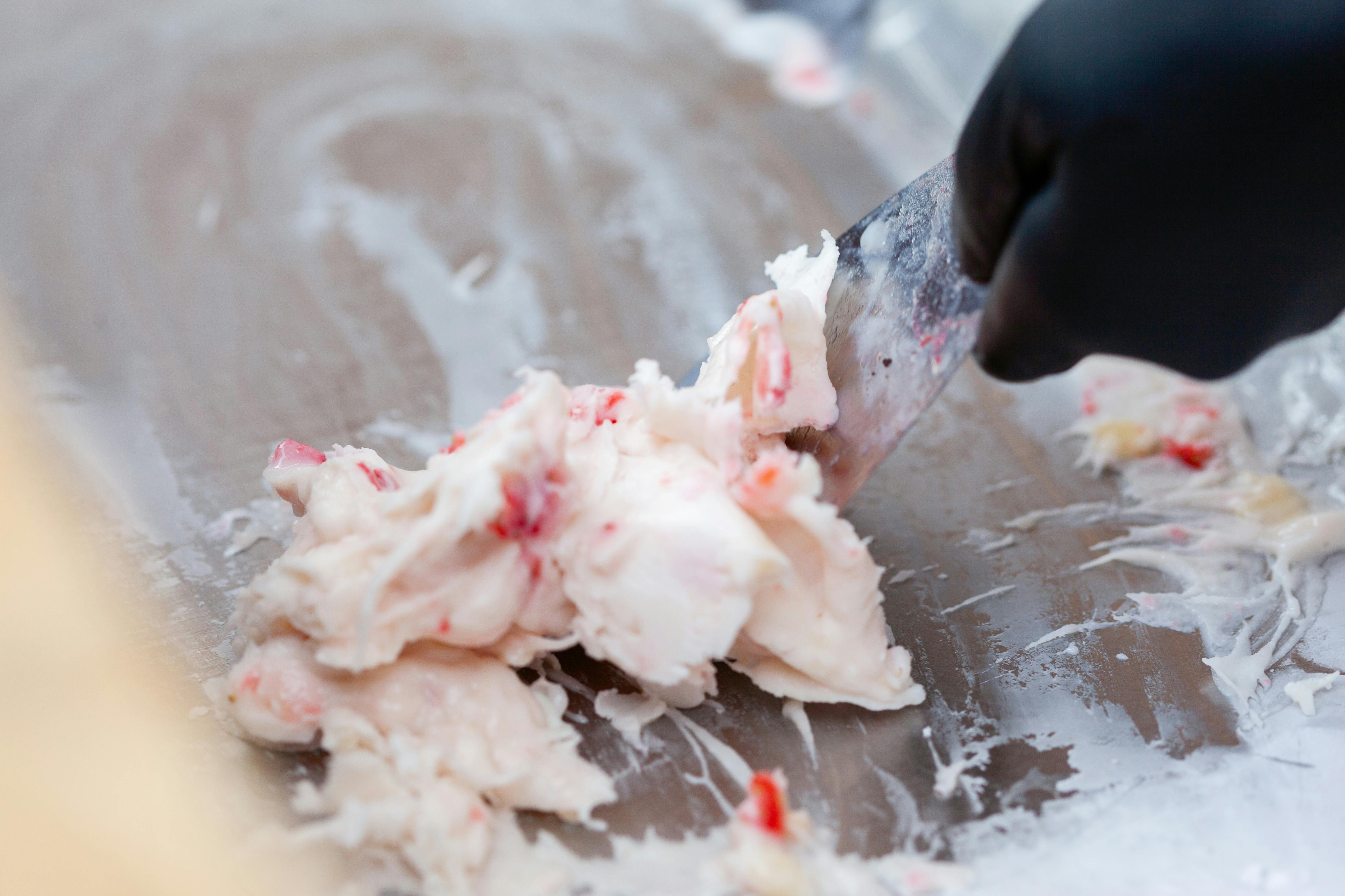Close-up of hand mixing creamy ice cream with strawberries on a cold metal surface.