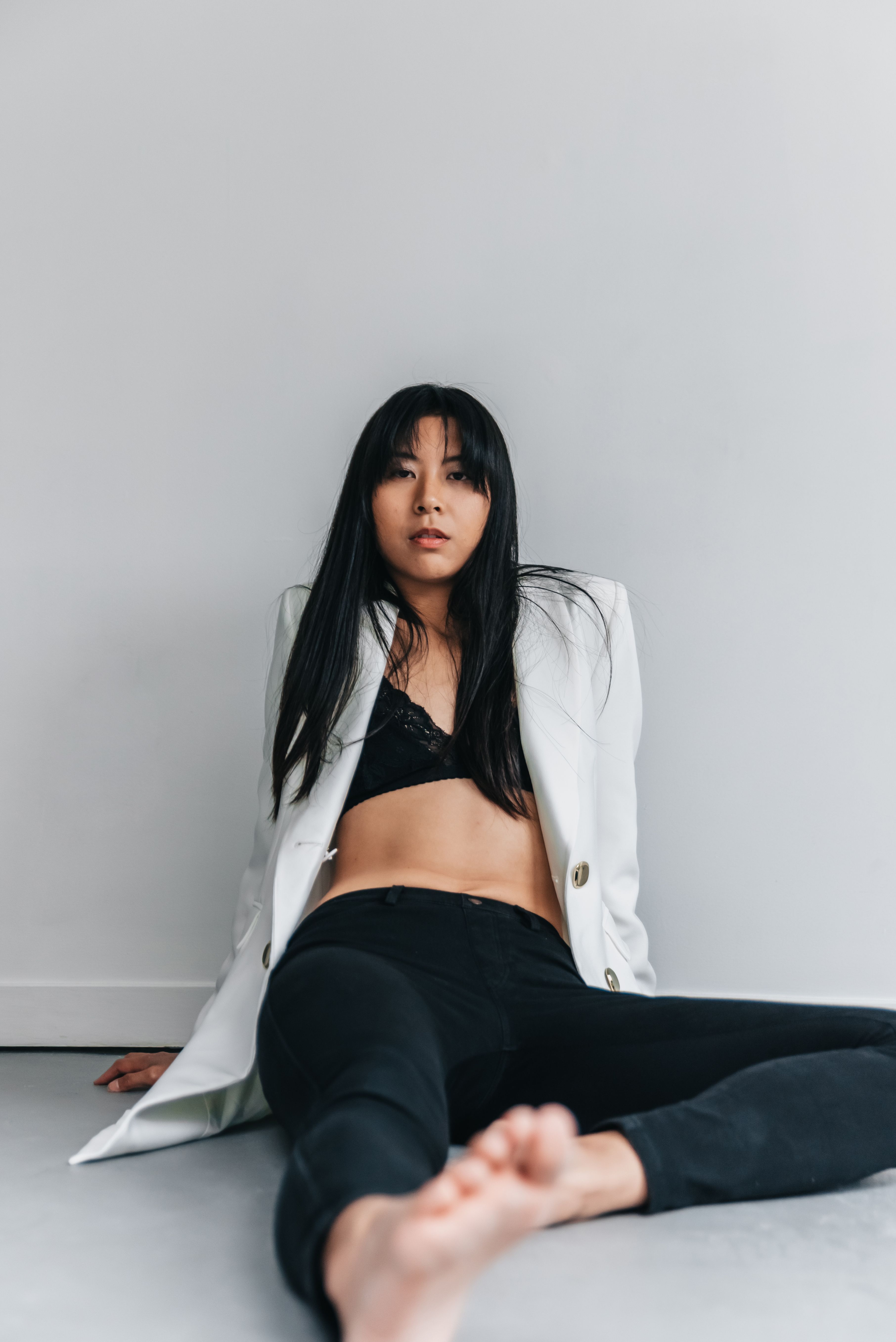 Elegant studio shot of an Asian woman in a white blazer and black bralette, sitting barefoot against a white backdrop.