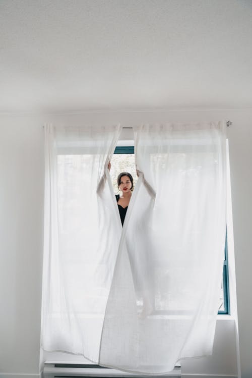 Free Woman Standing on the Window Holding the White Curtains  Stock Photo