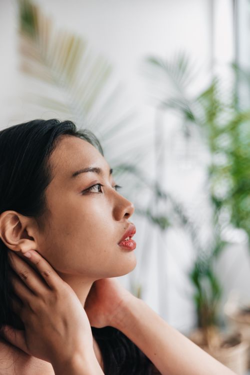 Free Portrait of an Asian woman posing confidently indoors with blurred tropical plants in the background. Stock Photo