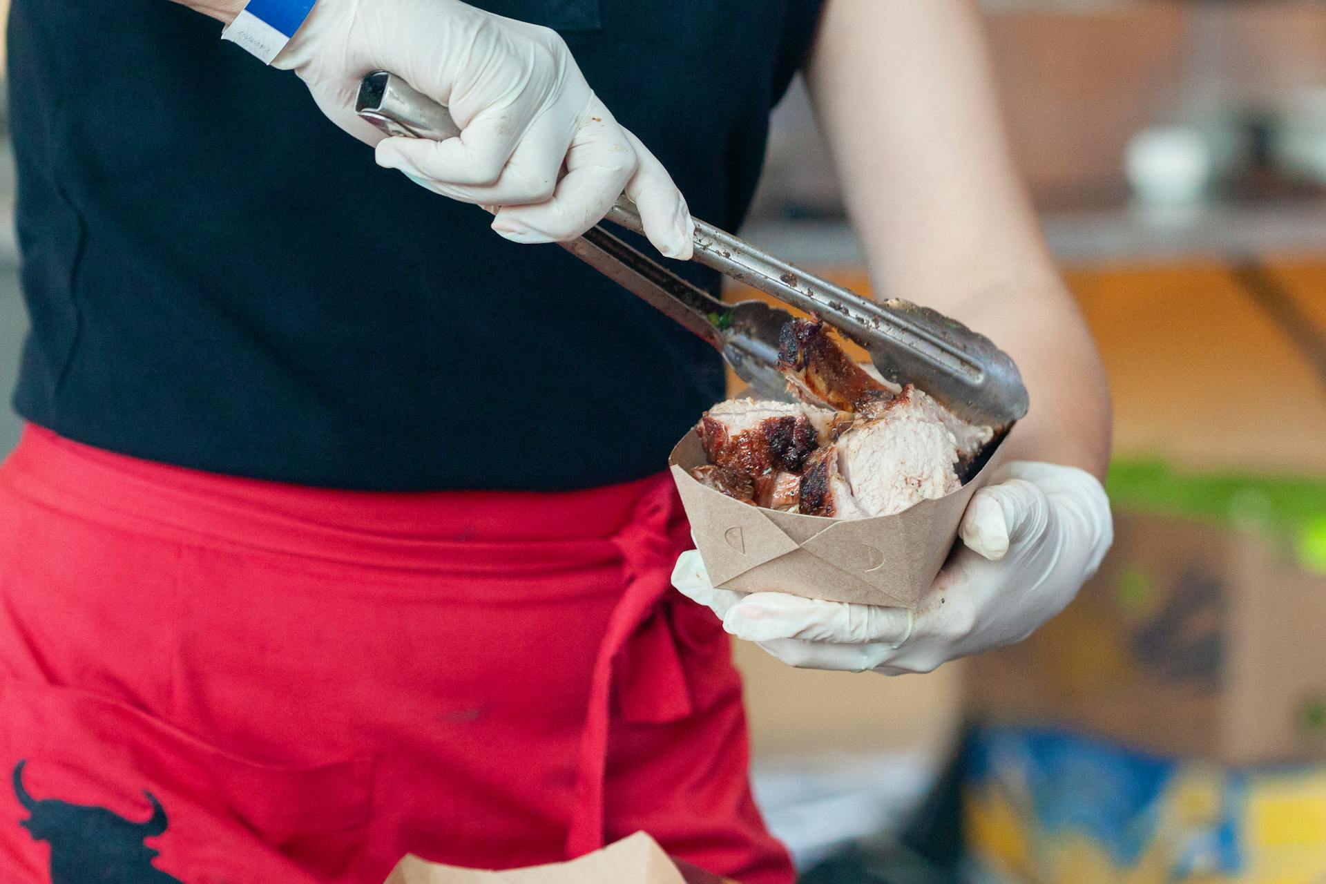 Gloved chef serving freshly grilled meat with tongs into a paper food tray at a food event.