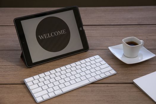 Stylish office setup with a tablet displaying 'WELCOME', a keyboard, and coffee on a wooden table.